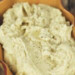 A serving dish filled with creamy Crock Pot Mashed Potatoes, topped with a sprinkle of black pepper, sits on a wooden surface.
