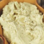 A serving dish filled with creamy Crock Pot Mashed Potatoes, topped with a sprinkle of black pepper, sits on a wooden surface.