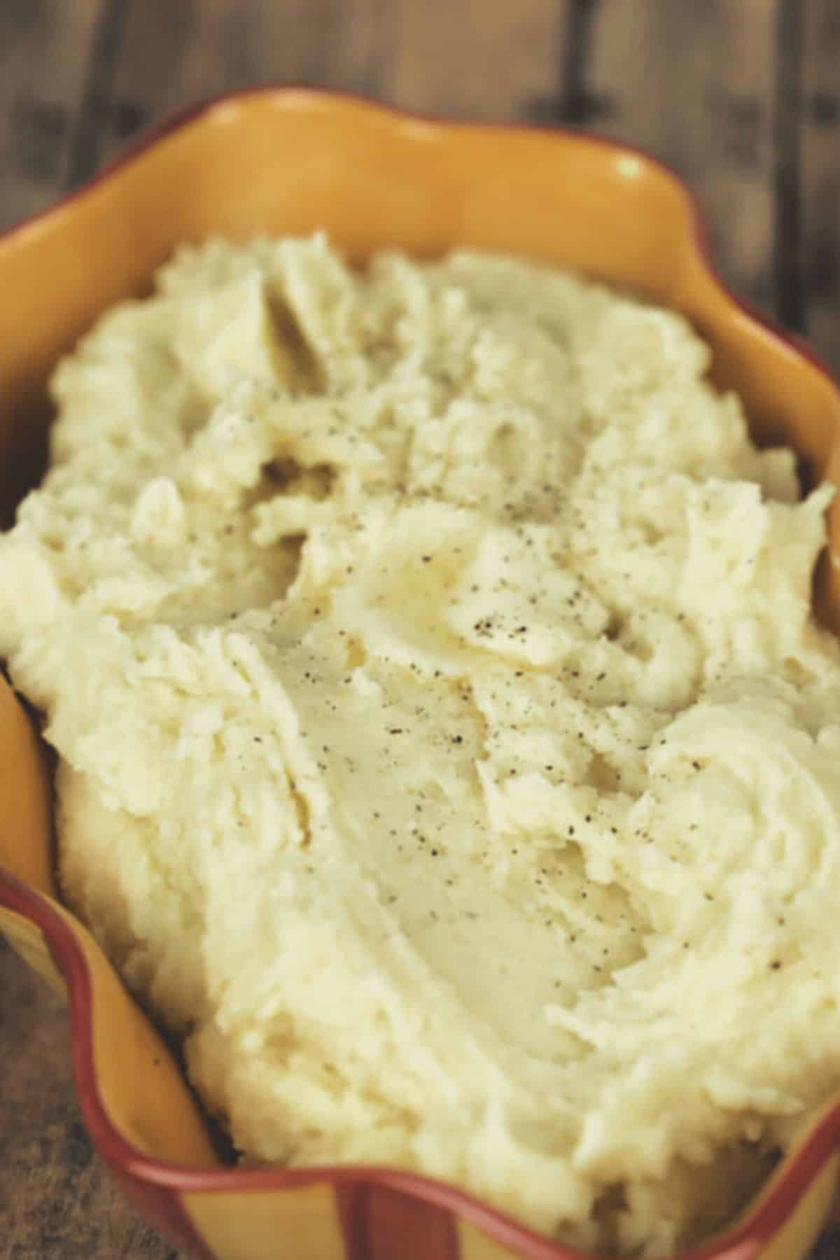 A serving dish filled with creamy Crock Pot Mashed Potatoes, topped with a sprinkle of black pepper, sits on a wooden surface.