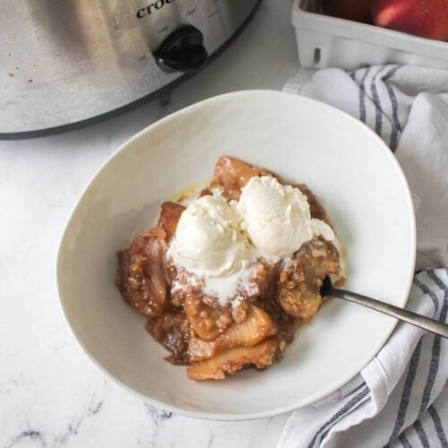 A bowl of crock pot peach cobbler topped with two scoops of vanilla ice cream sits next to a slow cooker and a box of fresh peaches on a white kitchen counter.