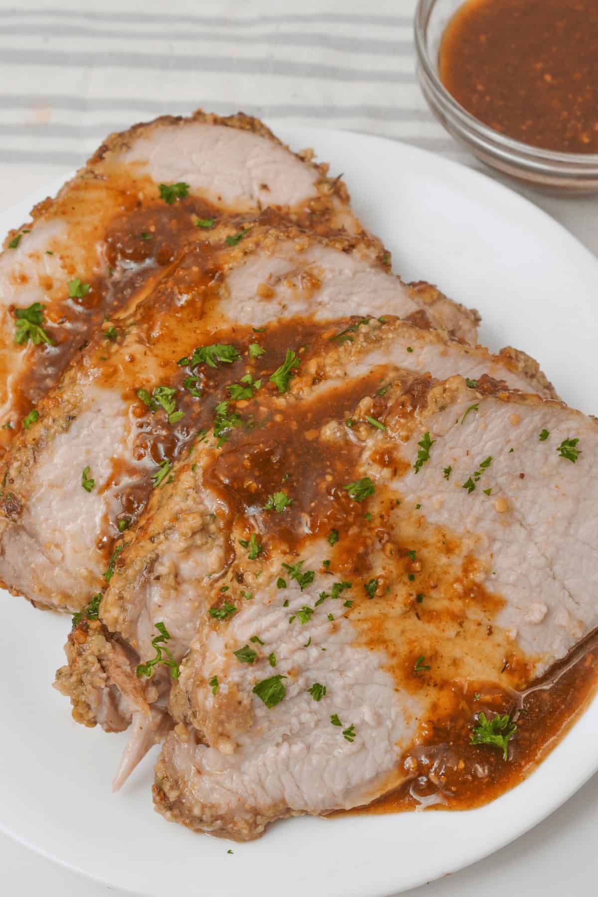 Slices of roasted crock pot pork tenderloin topped with sauce and garnished with herbs on a white plate. Nearby is a small bowl containing additional sauce.