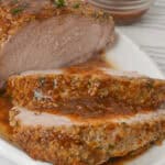 Sliced crock pot pork tenderloin with an herb crust on a white plate, accompanied by a small bowl of sauce.