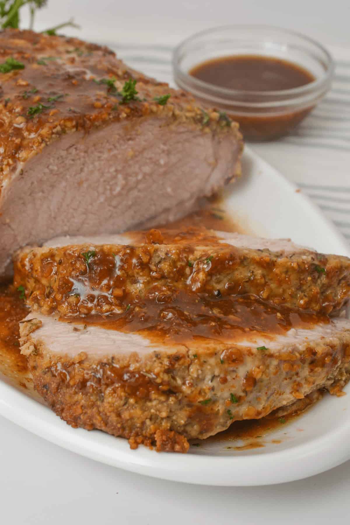 Sliced crock pot pork tenderloin with an herb crust on a white plate, accompanied by a small bowl of sauce.