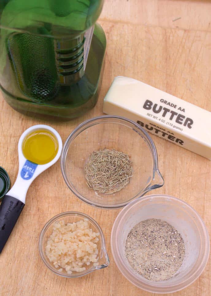 A bottle of oil, a stick of butter, a tablespoon of oil, dried rosemary, minced garlic, and ground pepper sit on a wooden surface next to a pile of Crock Pot Red Potatoes.