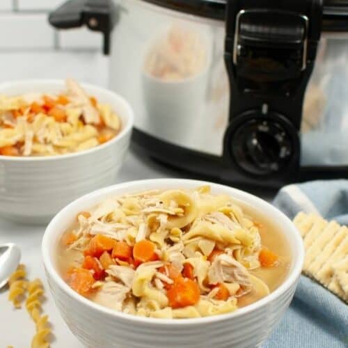 A white bowl of turkey soup with a crock pot beside it.