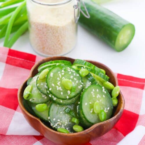 A wooden bowl of Cucumber Edamame Salad topped with sesame seeds sits on a red and white checkered cloth, with cucumber, green onions, and a jar of sesame seeds in the background.