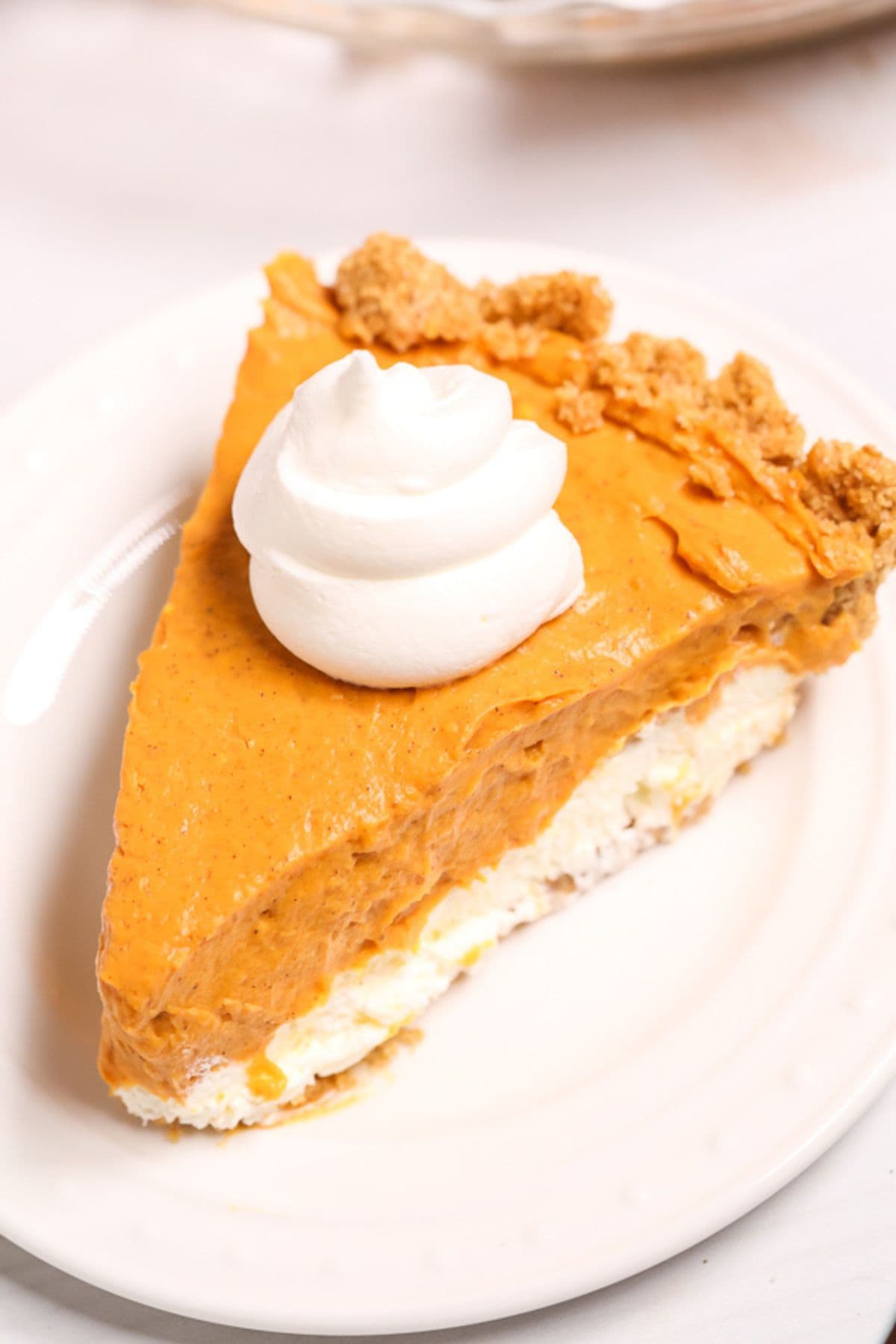 A slice of cream cheese pumpkin pie with a whipped cream topping and crumb garnish is served on a white plate.