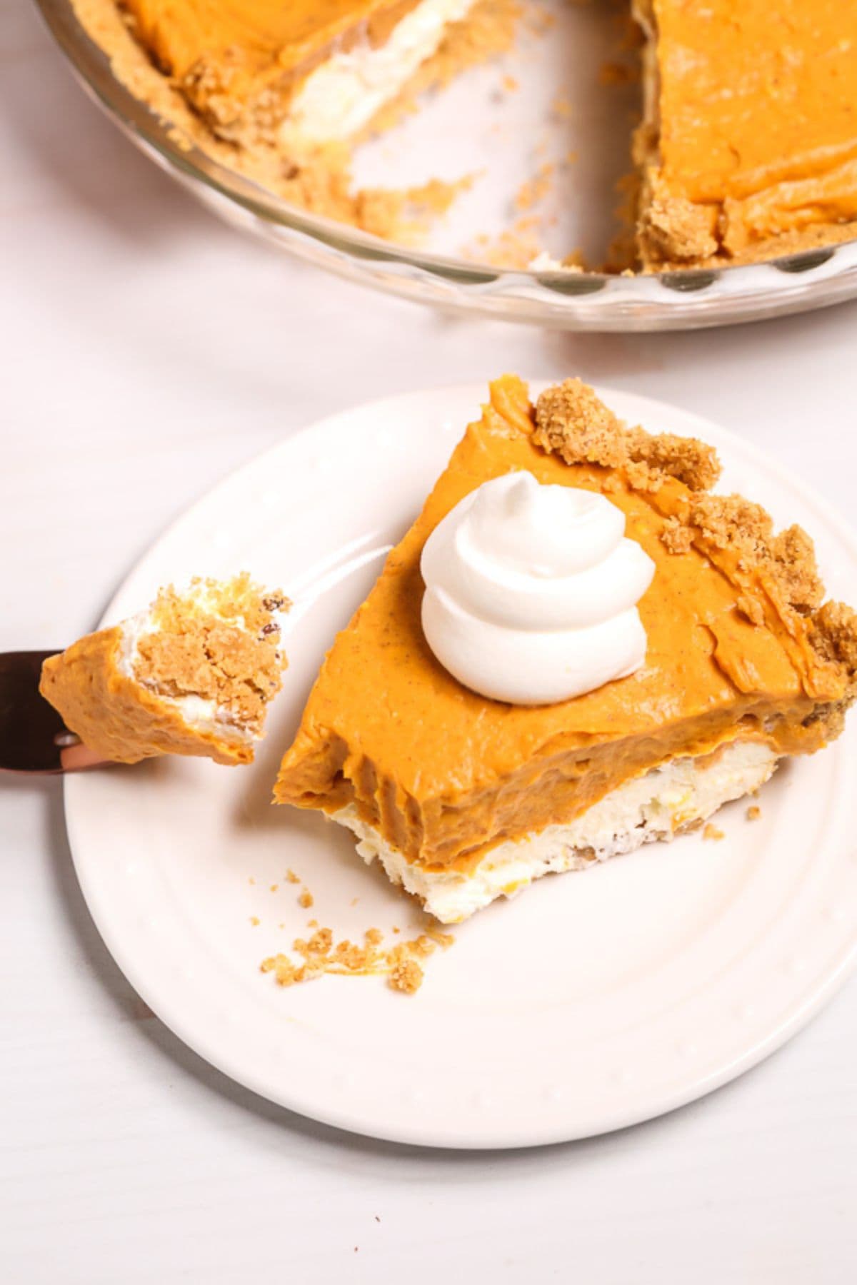 A slice of dessert with whipped cream on top sits on a white plate, with a fork holding a bite. The rest of the pie is in the background.