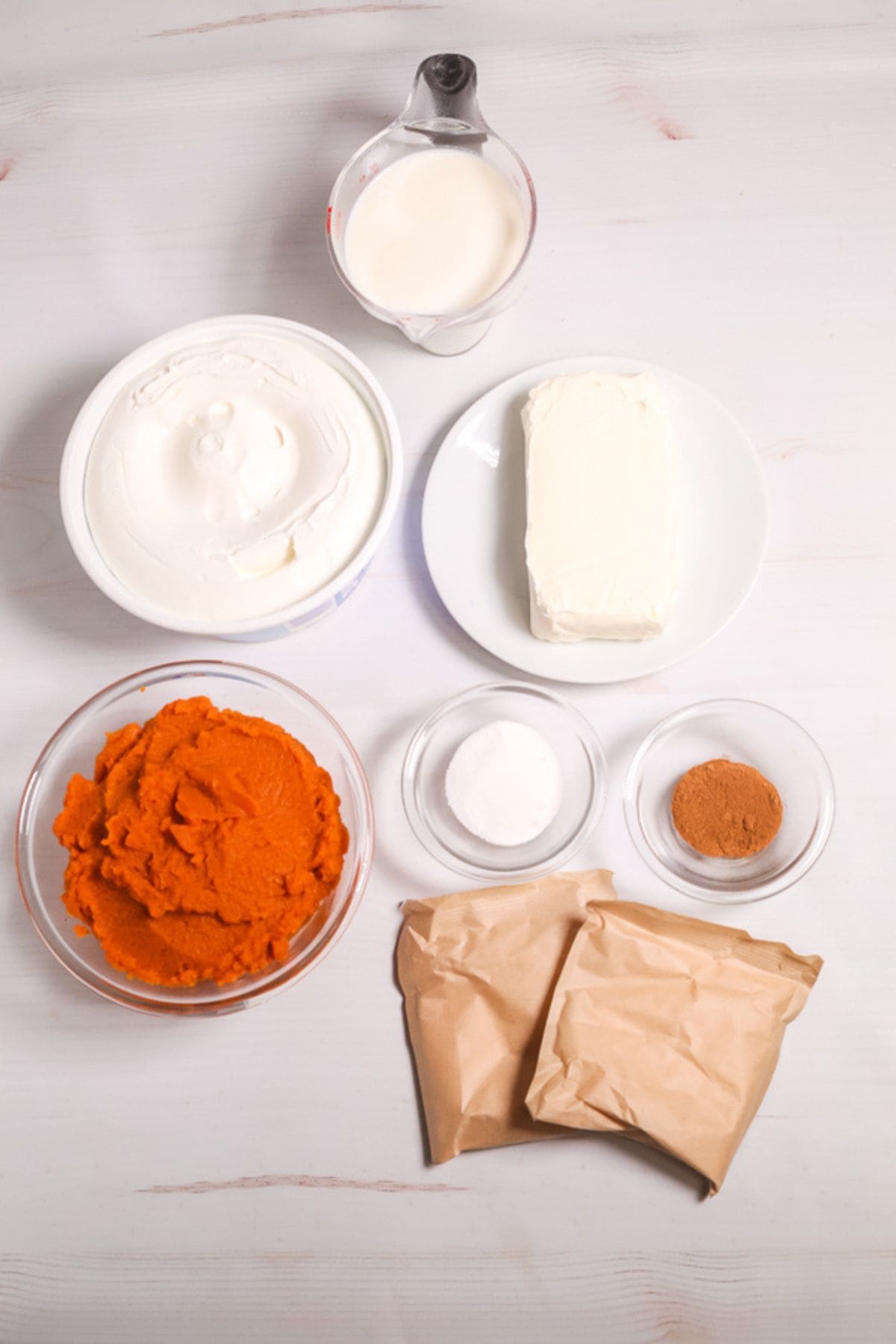 Ingredients on a white surface, featuring whipped topping, cream cheese, pumpkin puree, milk, sugar, cinnamon, and two packets in brown paper bags.