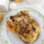 A Dutch Oven Roasted Chicken with herbs sits on a decorative plate; garlic, onion, fresh herbs, and a pot of broth are in the background.