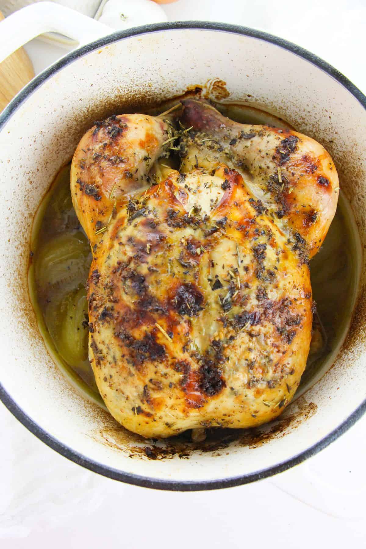 Roasted  with herbs and browned skin sits in a white pot surrounded by savory juices and tender cooked onions.