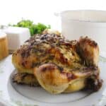 A Dutch Oven Roasted Chicken seasoned with herbs sits on a decorative plate, with a white pot and greenery in the background.