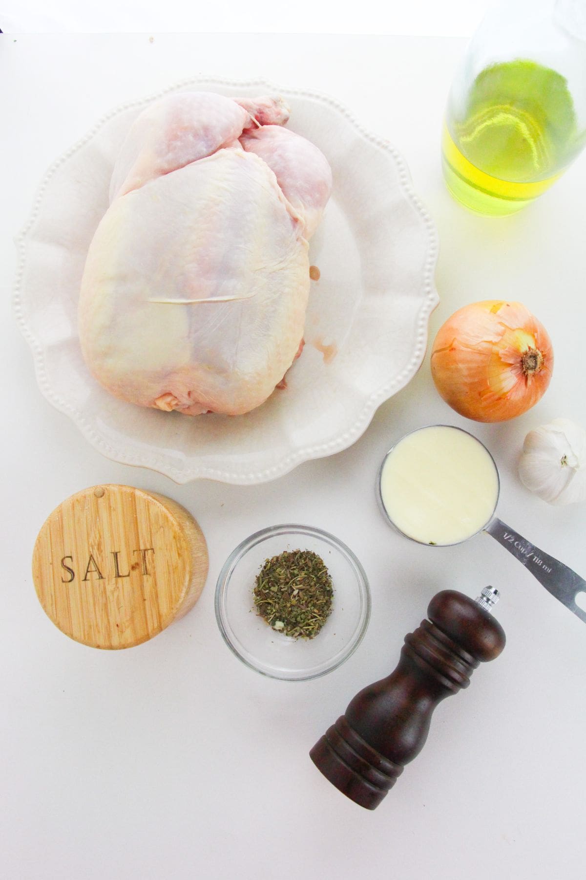 A whole raw chicken on a plate is ready to become Dutch Oven Roasted Chicken, surrounded by olive oil, onion, garlic, salt, dried herbs, black pepper grinder, and a cup of creamy liquid.