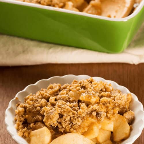 A bowl of easy apple crumble with a crumbly topping is in the foreground. A baking dish with more apple crisp and a wooden spoon is in the background, promising effortless indulgence.