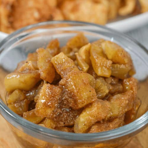 A clear bowl filled with spiced apple slices paired with an easy apple dip sits in focus, while a plate of crispy snacks is blurred in the background.