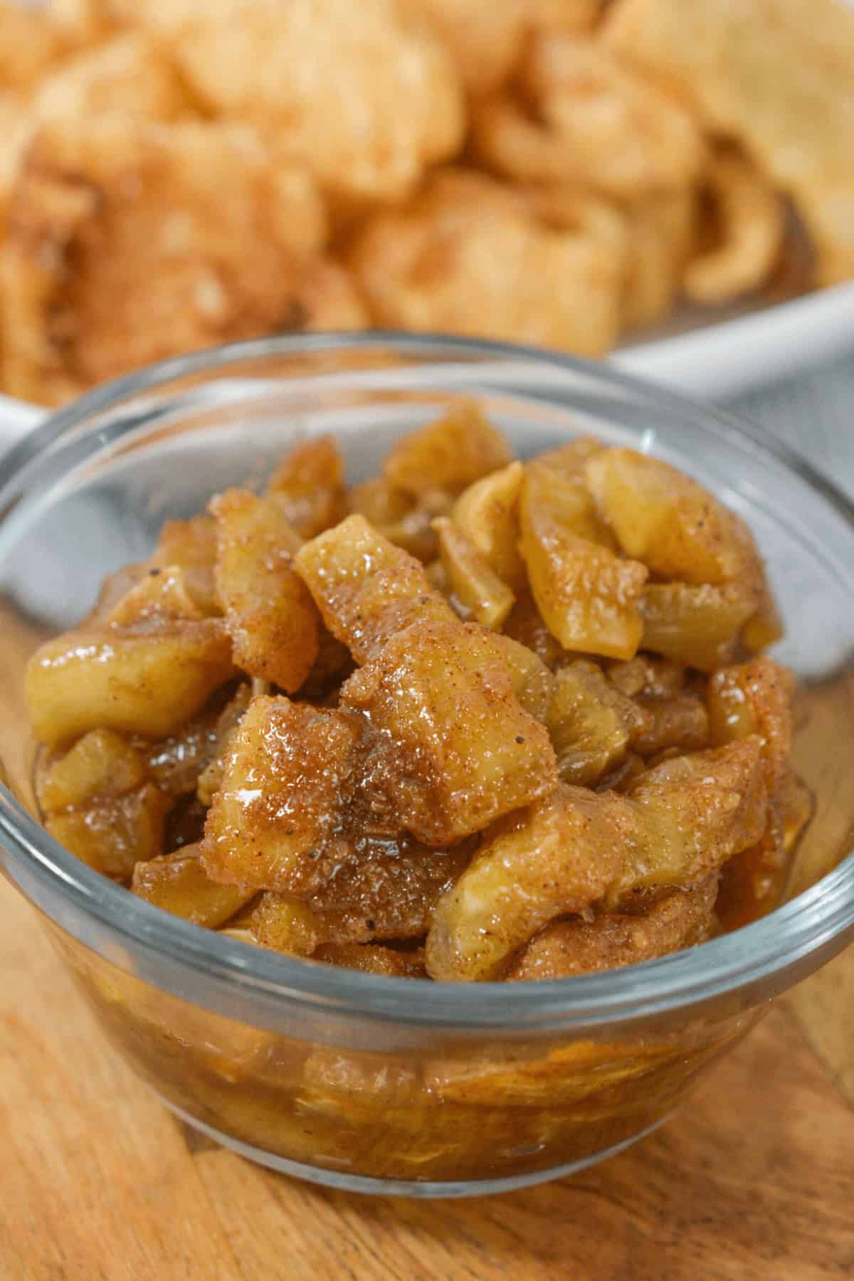 A clear bowl filled with spiced apple slices paired with an easy apple dip sits in focus, while a plate of crispy snacks is blurred in the background.