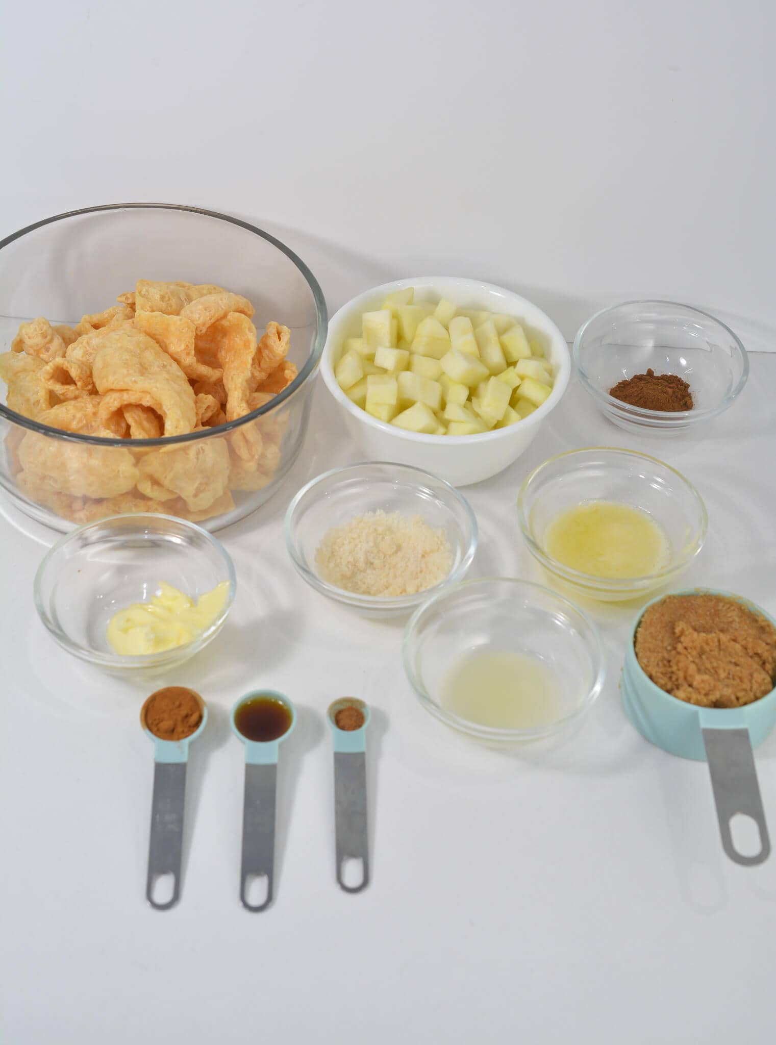 Ingredients for an easy apple dip on a table: pork rinds, diced apples, nutmeg, butter, sweetener, lemon juice, cinnamon, vanilla extract, almond flour, and brown sugar substitute arranged in various bowls and spoons.