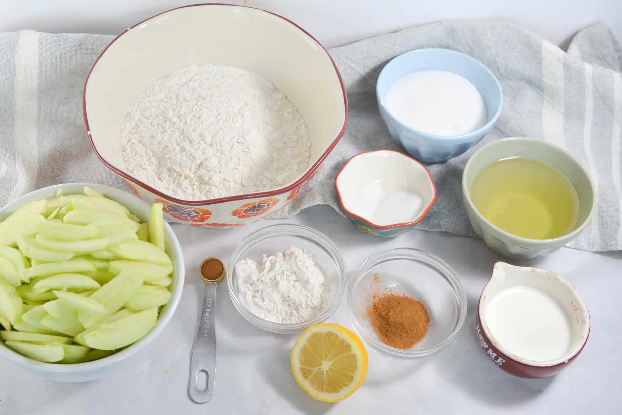 A neatly arranged selection of easy apple pie baking ingredients, including sliced apples, flour, sugar, oil, milk, lemon zest, cinnamon, and other essentials on a white surface with a striped cloth background.