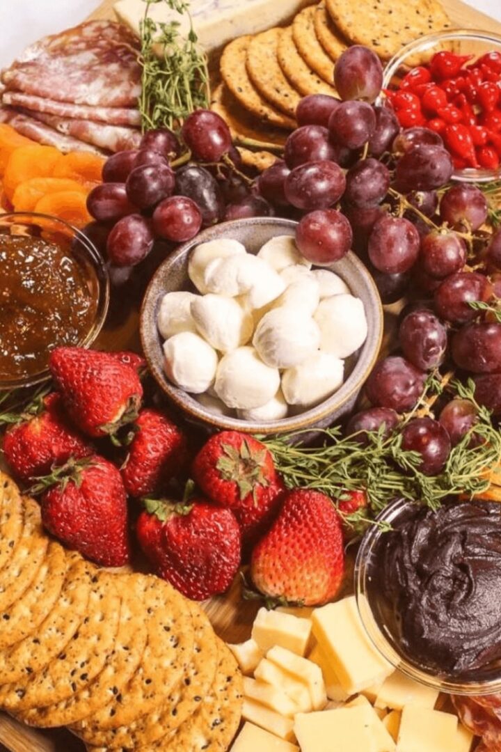 An easy charcuterie board featuring crackers, grapes, strawberries, mozzarella balls, salami, dried apricots, red berries, jam, and cheese cubes.