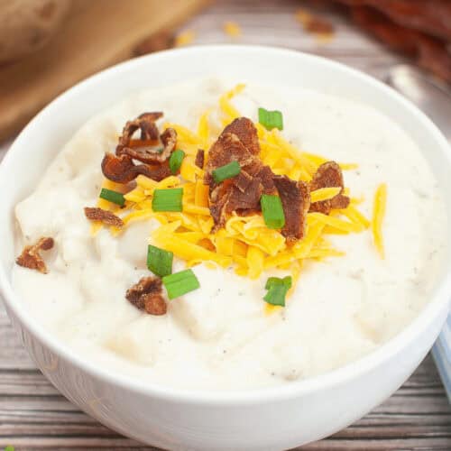 A bowl of creamy potato soup topped with shredded cheddar cheese, crumbled bacon, and chopped green onions, presented as a delectable Crock Pot Potato Soup recipe on a wooden surface.