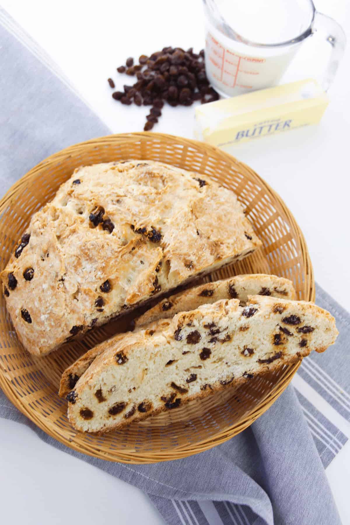 A loaf on a wicker plate with one slice cut. Nearby, a measuring cup with milk, butter, and raisins sits on a gray cloth—a comforting scene reminiscent of making easy Irish soda bread.
