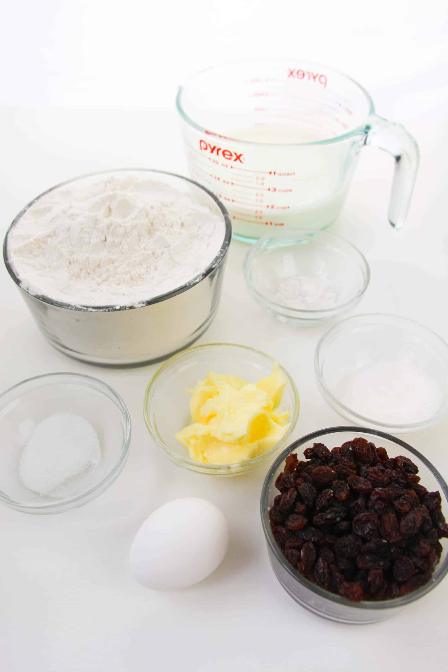 Various baking ingredients on a counter: a measuring cup, flour, butter, raisins, an egg, and small bowls containing sugar, baking powder, and salt.