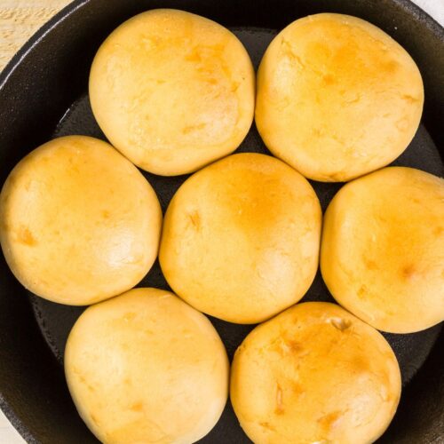 Seven golden bread arranged in a cast iron skillet, partially covered by a white and red kitchen towel.