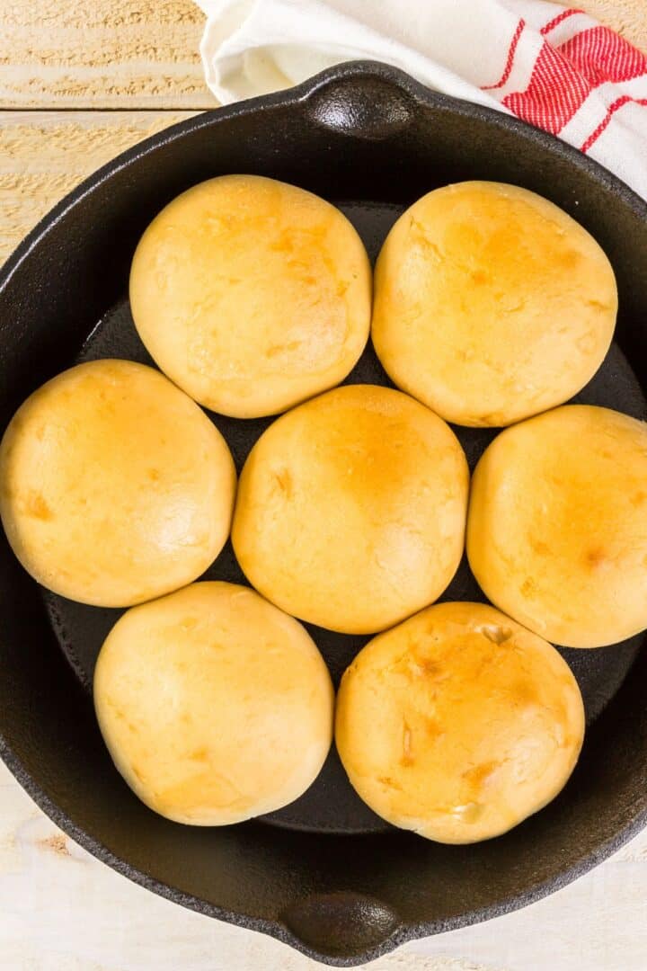 Seven golden bread arranged in a cast iron skillet, partially covered by a white and red kitchen towel.