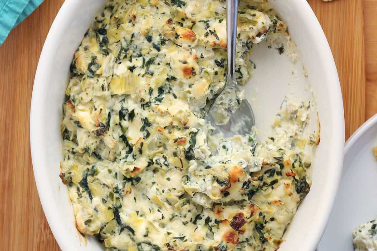 A white oval dish filled with baked spinach and artichoke dip, with a serving spoon and a portion removed, showing the creamy texture—a perfect choice for easy appetizers at your next gathering.