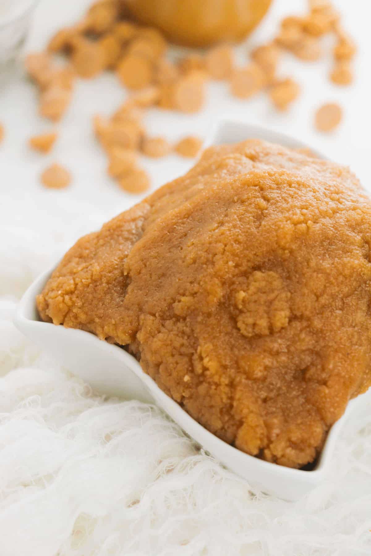 A mound of brown, edible peanut butter cookie dough sits in a small, white square dish, surrounded by scattered caramel bits on a white textured surface.