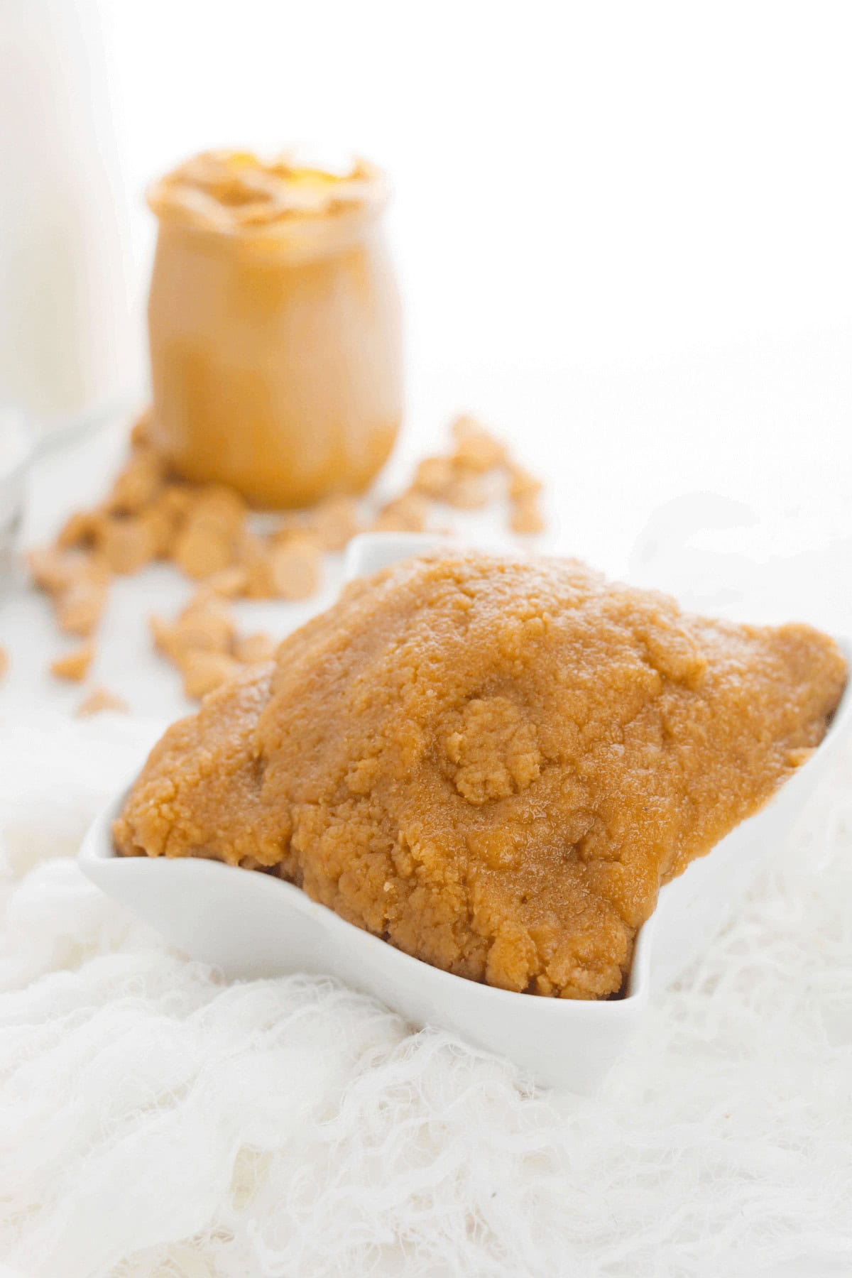 A small white dish filled with thick, brown edible peanut butter cookie dough rests atop a textured white surface. In the background, a jar of peanut butter and some scattered crumbs are visible.