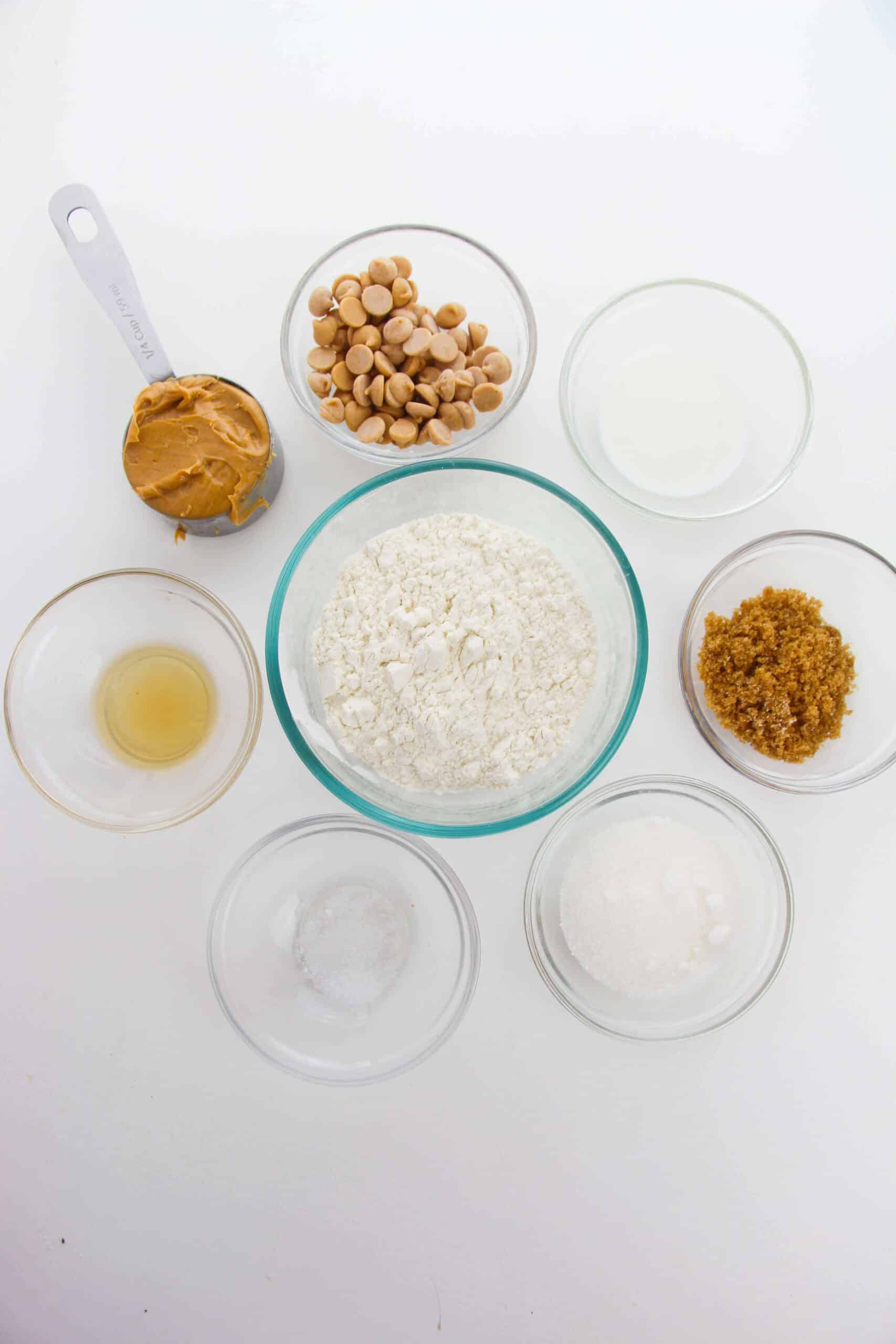 An assortment of edible peanut butter cookie dough ingredients in bowls: flour, peanut butter, butterscotch chips, brown sugar, white sugar, liquid, salt, and vanilla extract arranged on a white surface.