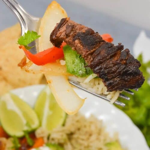 A fork holding grilled steak, onion, red pepper, and green pepper above a vibrant dish with avocado, sour cream, and lime wedges.