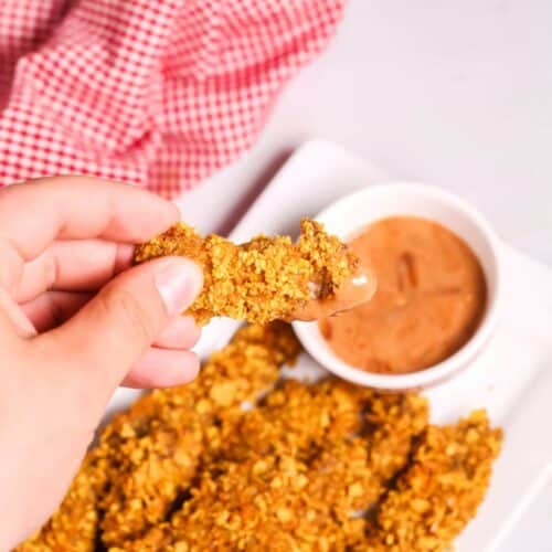A hand holds a finger steak dipped in sauce, with more crispy chicken strips on a white plate beside a bowl of dipping sauce. A red and white checkered cloth adds a cozy background touch.