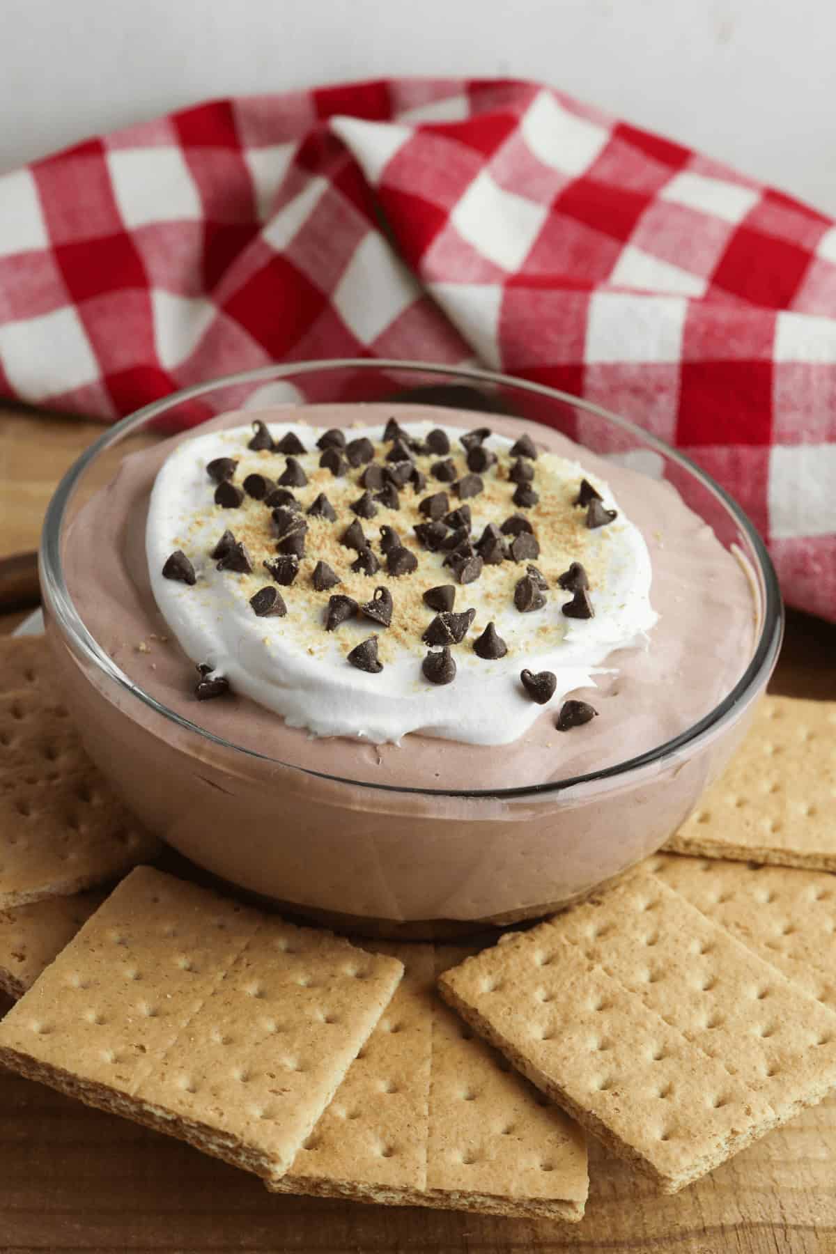 A bowl of with whipped topping and chocolate chips is surrounded by graham crackers on a wooden surface, with a red-checkered cloth in the background.