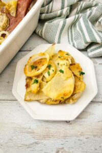 A serving of Potato Bake garnished with herbs on a white plate, next to a baking dish and a striped kitchen towel.