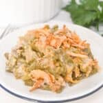 A serving of French’s Green Bean Casserole with crispy fried onions sits on a white plate, with the casserole dish and fresh parsley in the background.