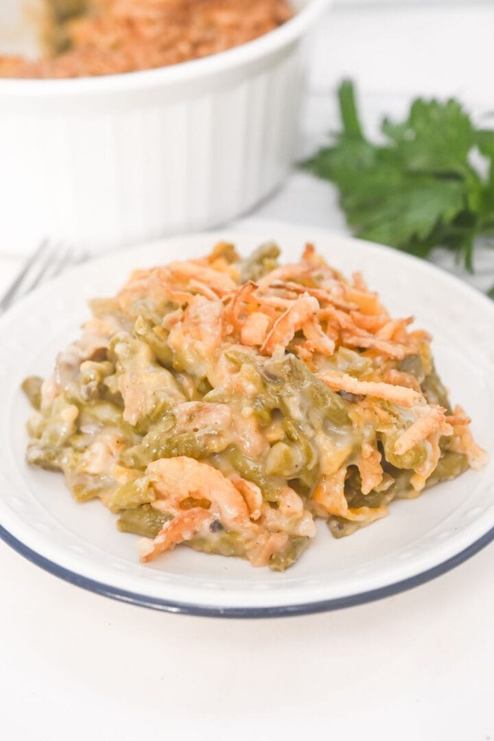 A serving of French’s Green Bean Casserole with crispy fried onions sits on a white plate, with the casserole dish and fresh parsley in the background.