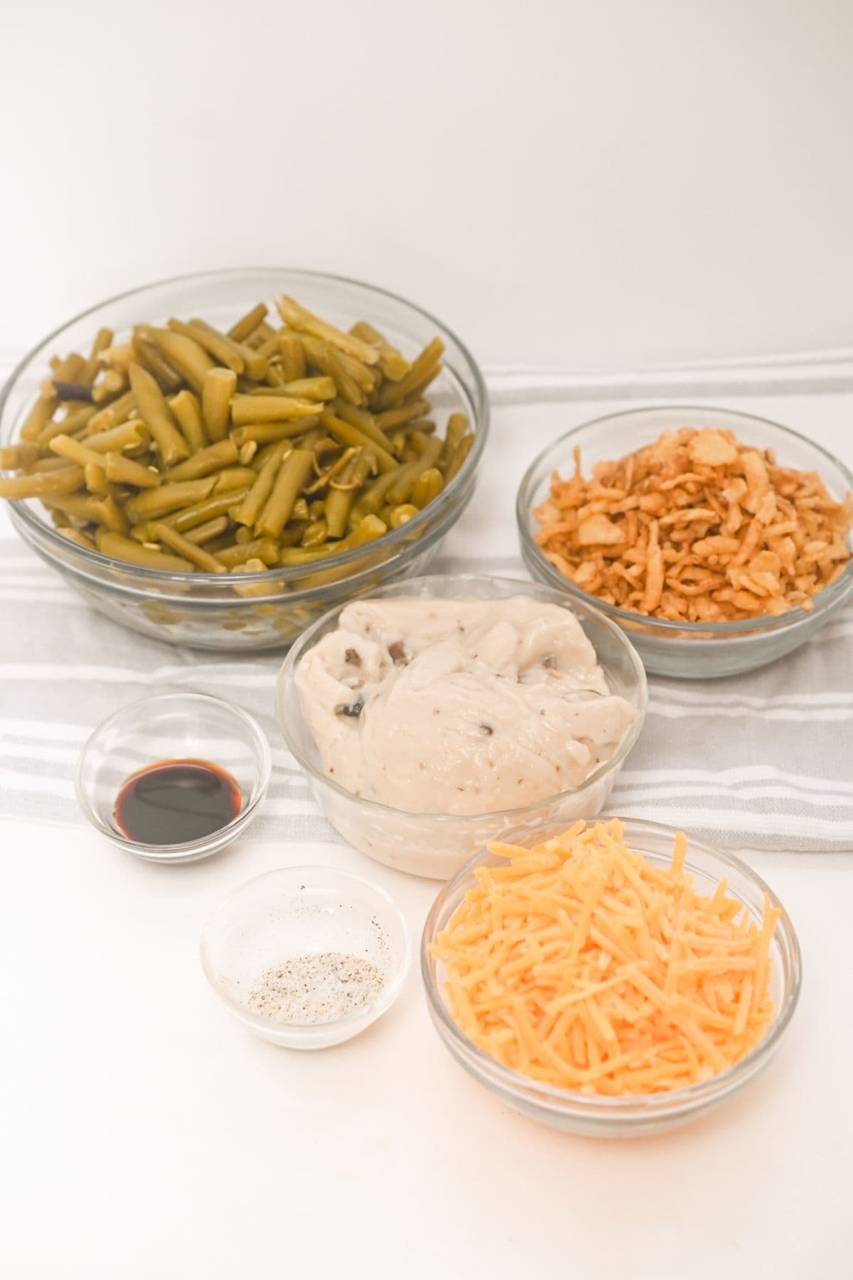 Five bowls canned vegetables, fried onions, cream of mushroom soup, shredded cheddar cheese, soy sauce, and salt and pepper—are arranged neatly on a striped towel.