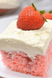 A slice of Strawberry Poke Cake topped with a thick layer of whipped cream and a whole strawberry placed on top, served on a white plate.