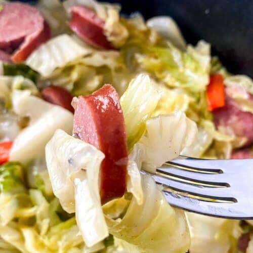 A fork holding a bite of Fried Cabbage and Sausage, with more cabbage, sausage, and vegetables visible in the background.