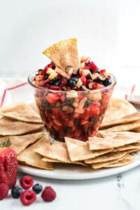A clear glass bowl filled with a delightful fruit salsa, featuring diced apples, strawberries, and blueberries, surrounded by cinnamon-sugar coated tortilla chips on a white plate.