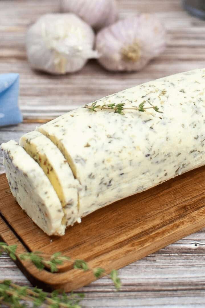 A garlic herb butter log with two slices cut, rests on a wooden board, garnished with fresh herbs. Garlic bulbs linger in the background.