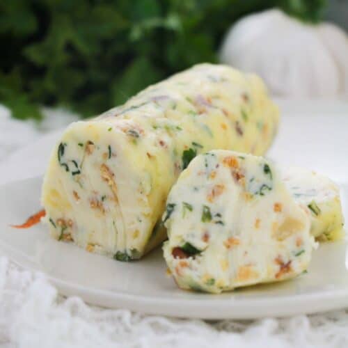 A log of Garlic Herb Steak Butter with herbs and vegetables is sliced on a white plate, with greenery in the blurred background.