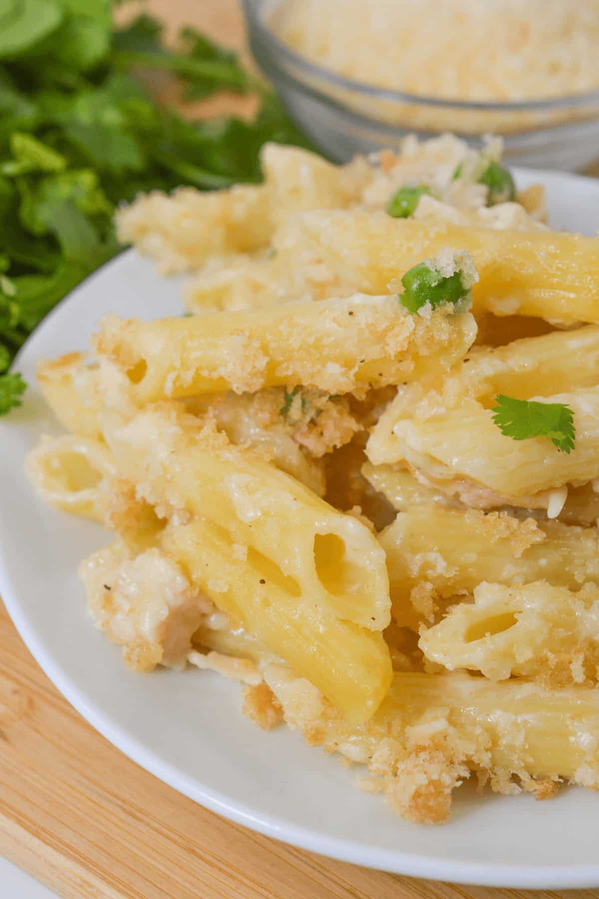 A plate of garlic Parmesan baked penne pasta with creamy sauce, tender chicken, and a sprinkle of parsley sits invitingly on a wooden board. In the background, a bowl of grated cheese awaits to enhance the savory delight.