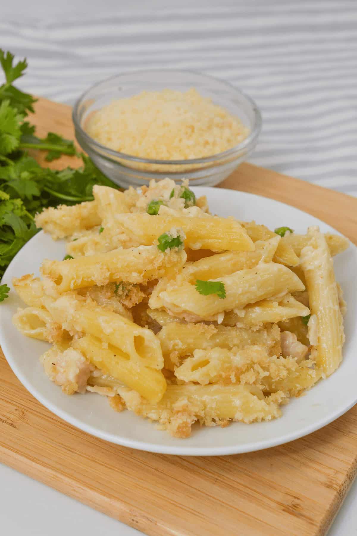 A plate of creamy garlic Parmesan penne pasta garnished with parsley and tender chicken pieces, placed on a wooden board. A small bowl of grated cheese is in the background.