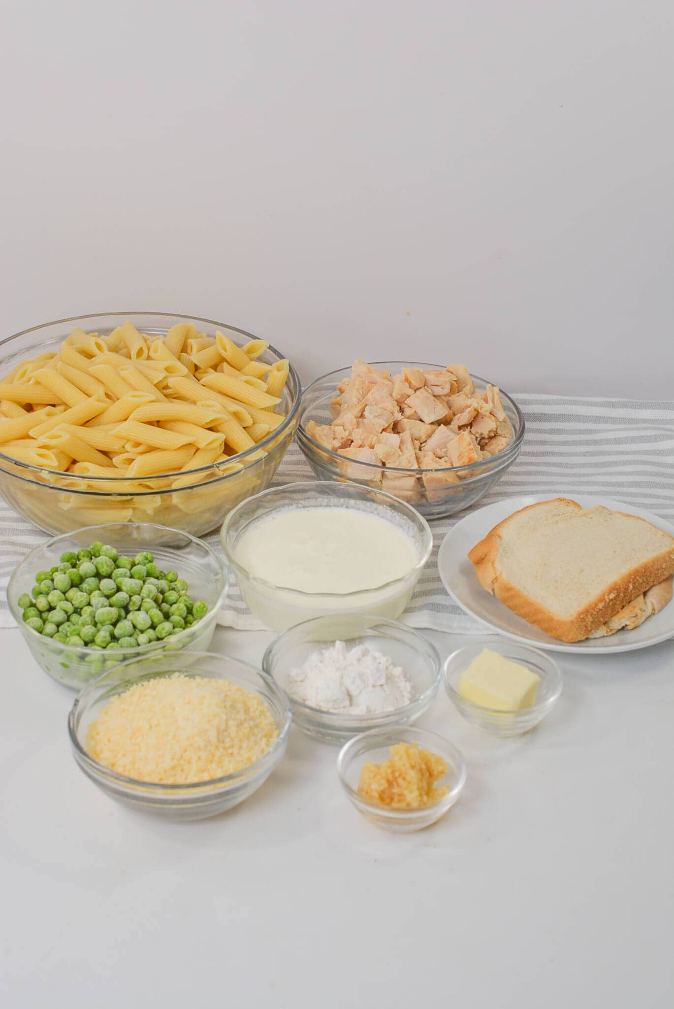 Ingredients for Garlic Parmesan Pasta with Chicken, including uncooked penne, diced chicken, peas, milk, bread, shredded cheese, butter, garlic, and flour in separate bowls on a striped cloth.