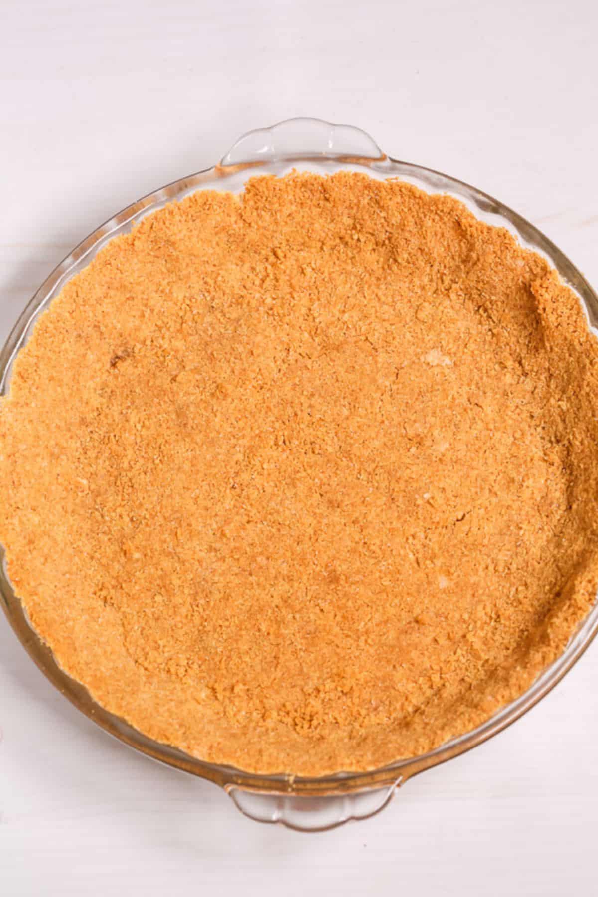 A glass pie dish filled with a baked, golden-brown Graham Cracker Pie Crust sits on a light-colored surface.