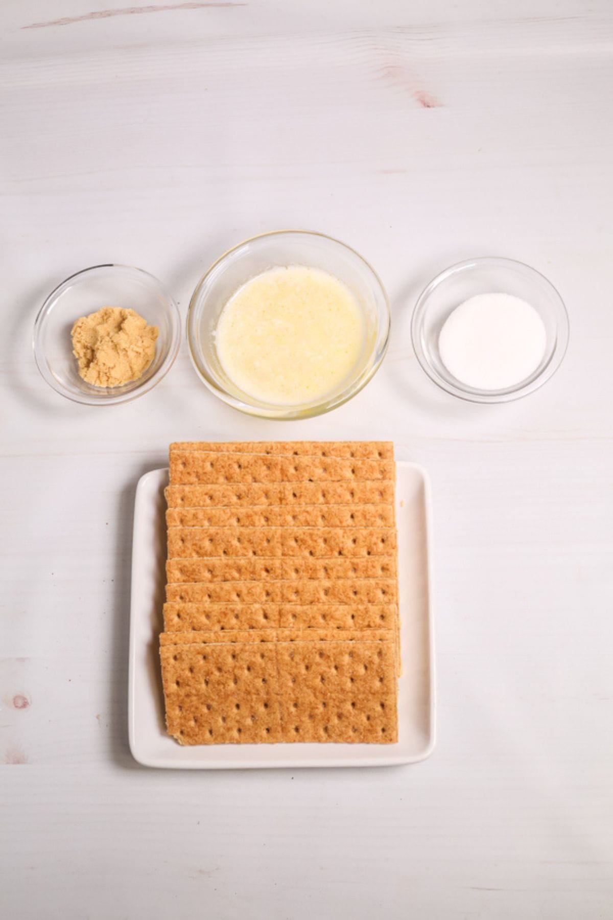 Rectangular graham crackers on a white plate, with three small bowls above containing brown sugar, melted butter, and granulated sugar—perfect ingredients for making a homemade Graham Cracker Pie Crust—all arranged neatly on a white surface.