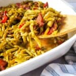 A wooden spoon lifts a serving of Green Beans with Bacon and Onion, mixed with tomato, from a white baking dish.